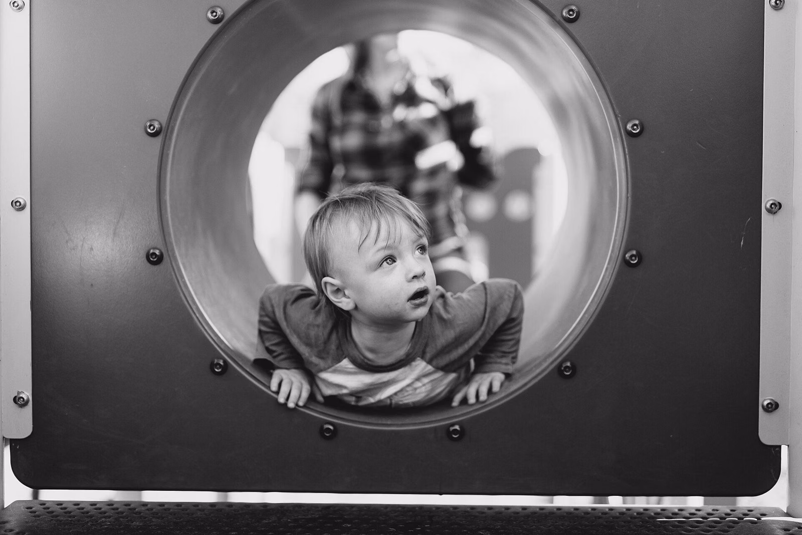A small child peeking out of a round tunnel on a playground structure, in black and white